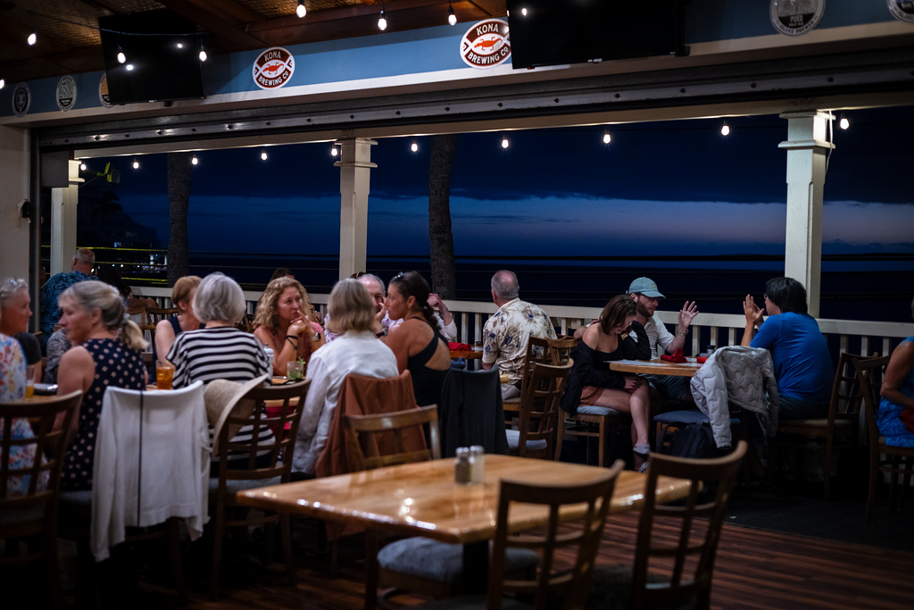 Diners at an open-air oceanfront restaurant in Kona at dusk, strung with lights