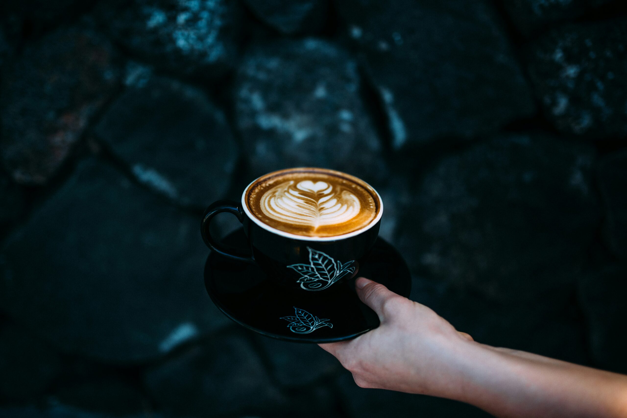 Hand holding a cup of coffee with rosetta latte art against a dark stone wall